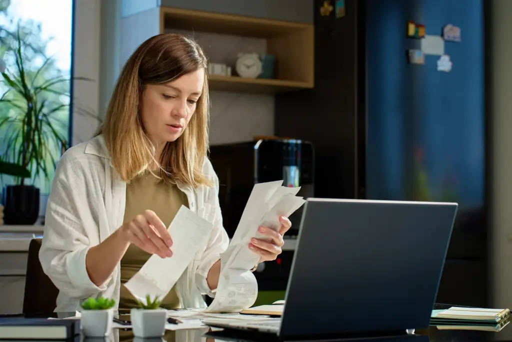 Self-employed IVAs: self-employed woman reviewing bills. Female organizing receipts, sitting at kitchen table with laptop and paper documents.